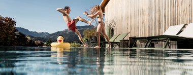 Two children joyfully jumping into a pool, with a rubber duck float, wooden house, and mountain landscape in the background on a sunny day. | © Kleinwalsertal Tourismus eGen • Photographer: Hannah Bichay