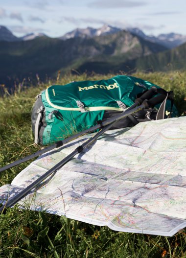 Hiking map, backpack, and trekking poles on a meadow with mountain views in the background. | © Kleinwalsertal Tourismus eGen • Photographer: Oliver Farys