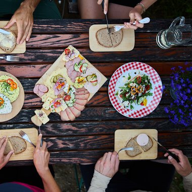 Top-down view of a table with cheese, meat, bread, and salad plates, surrounded by four people enjoying a meal. | © Kleinwalsertal Tourismus eGen • Photographer: Oliver Farys