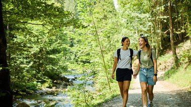 © Kleinwalsertal Tourismus eGen • Photographer: Oliver Farys Two young women walking and laughing along a lush forest trail by a river during summer 2024, surrounded by nature and sunshine. | © Kleinwalsertal Tourismus eGen • Photographer: Oliver Farys